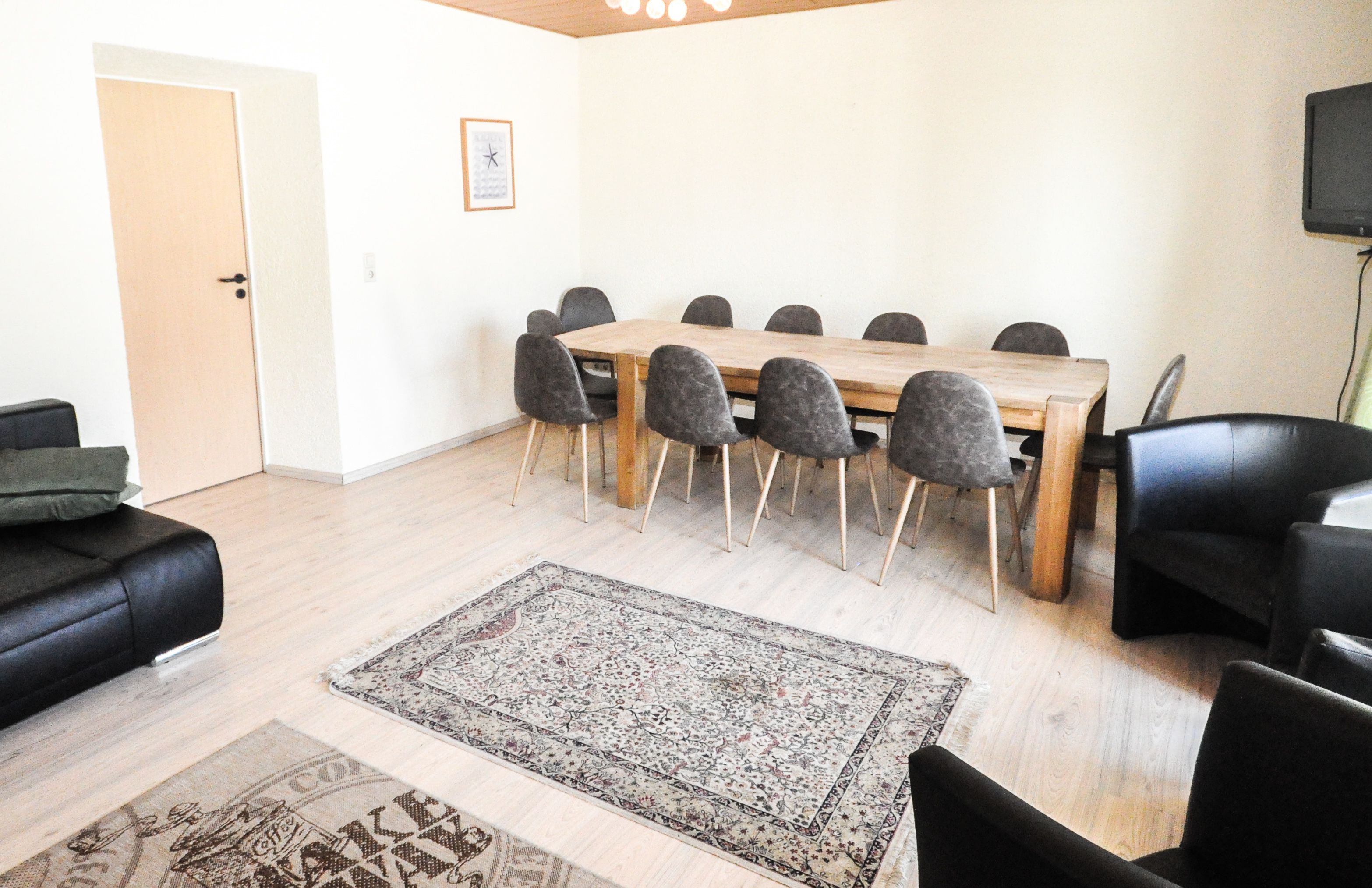 A dining area with a wooden table and eight chairs, a patterned rug on the floor, and a black leather sofa.