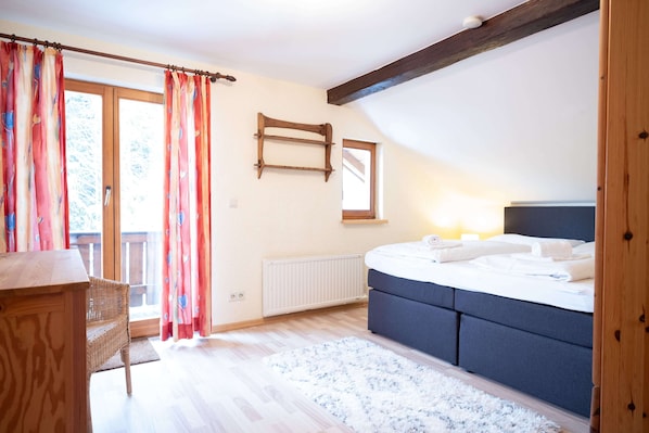A bedroom with a bed, a wooden dresser, a wicker chair, a radiator, and a window with curtains. - Lake View Lodges by we rent (Zell am See)