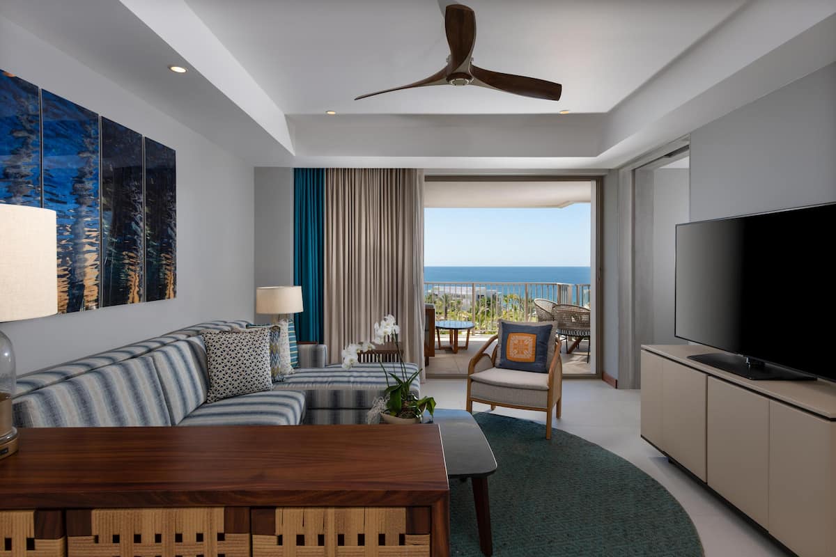 A modern hotel room with a balcony, a large flat-screen TV, a sofa, a dining table, and a ceiling fan.