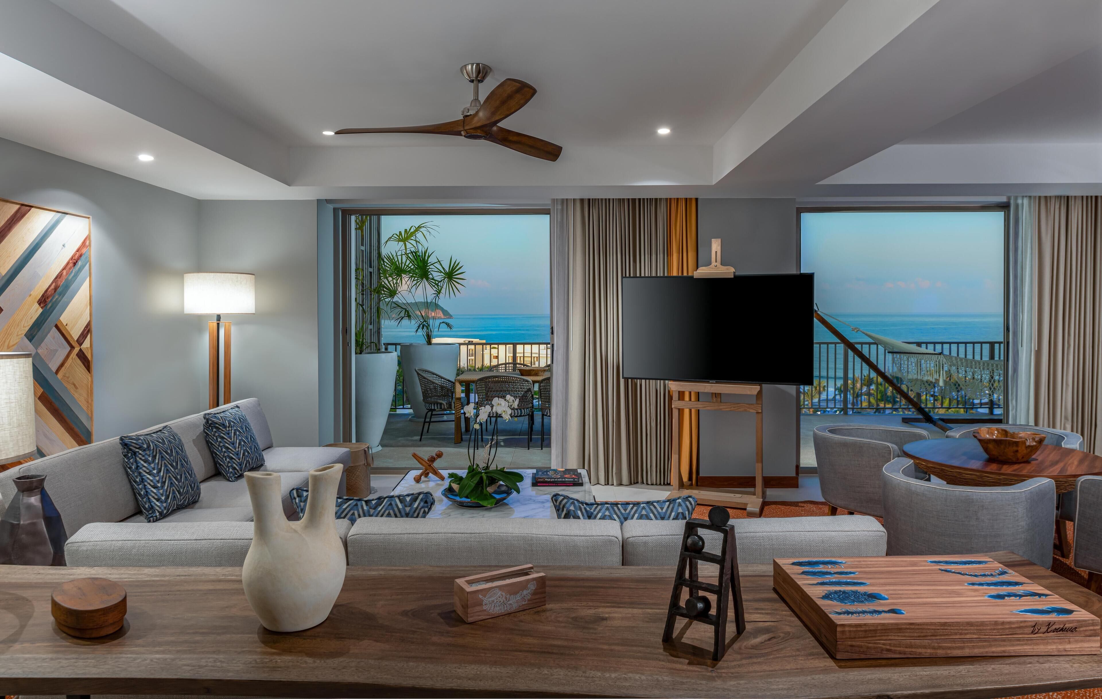 Una sala de estar moderna con sofá, mesa de centro y televisor, con vista a un balcón y al océano.