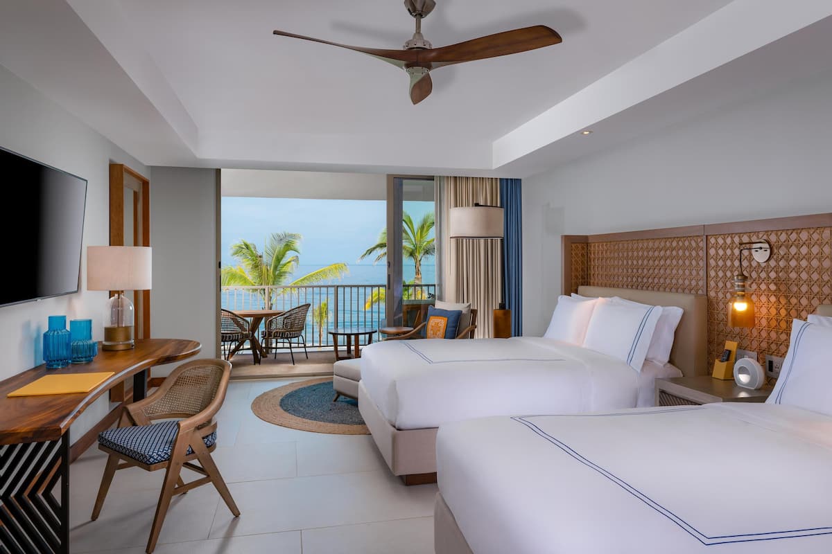 A hotel room with two beds, a desk, a TV, and a balcony with a view of palm trees and the ocean.