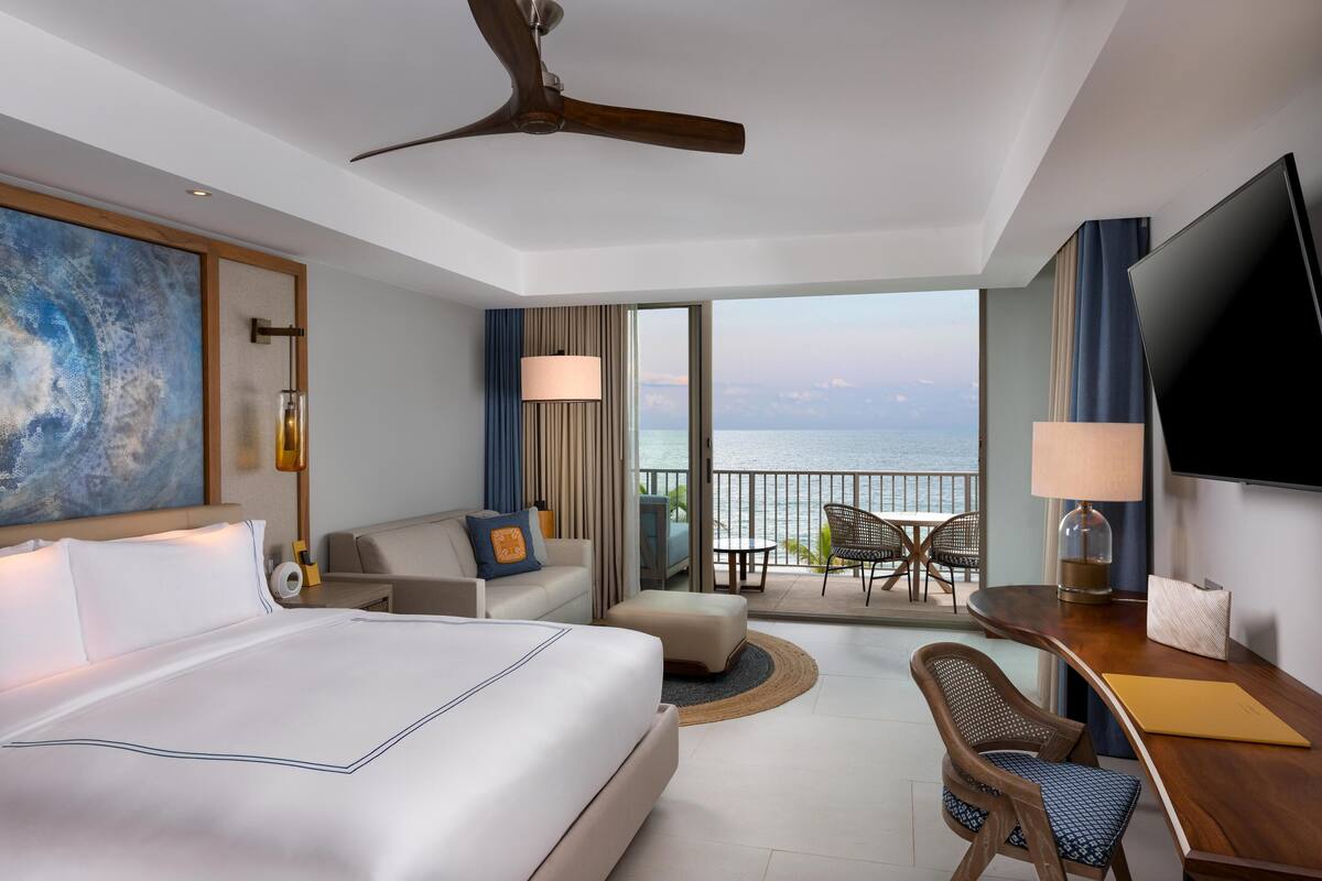 A hotel room with a bed, a desk, a chair, a TV, and a balcony with a view of the sea.