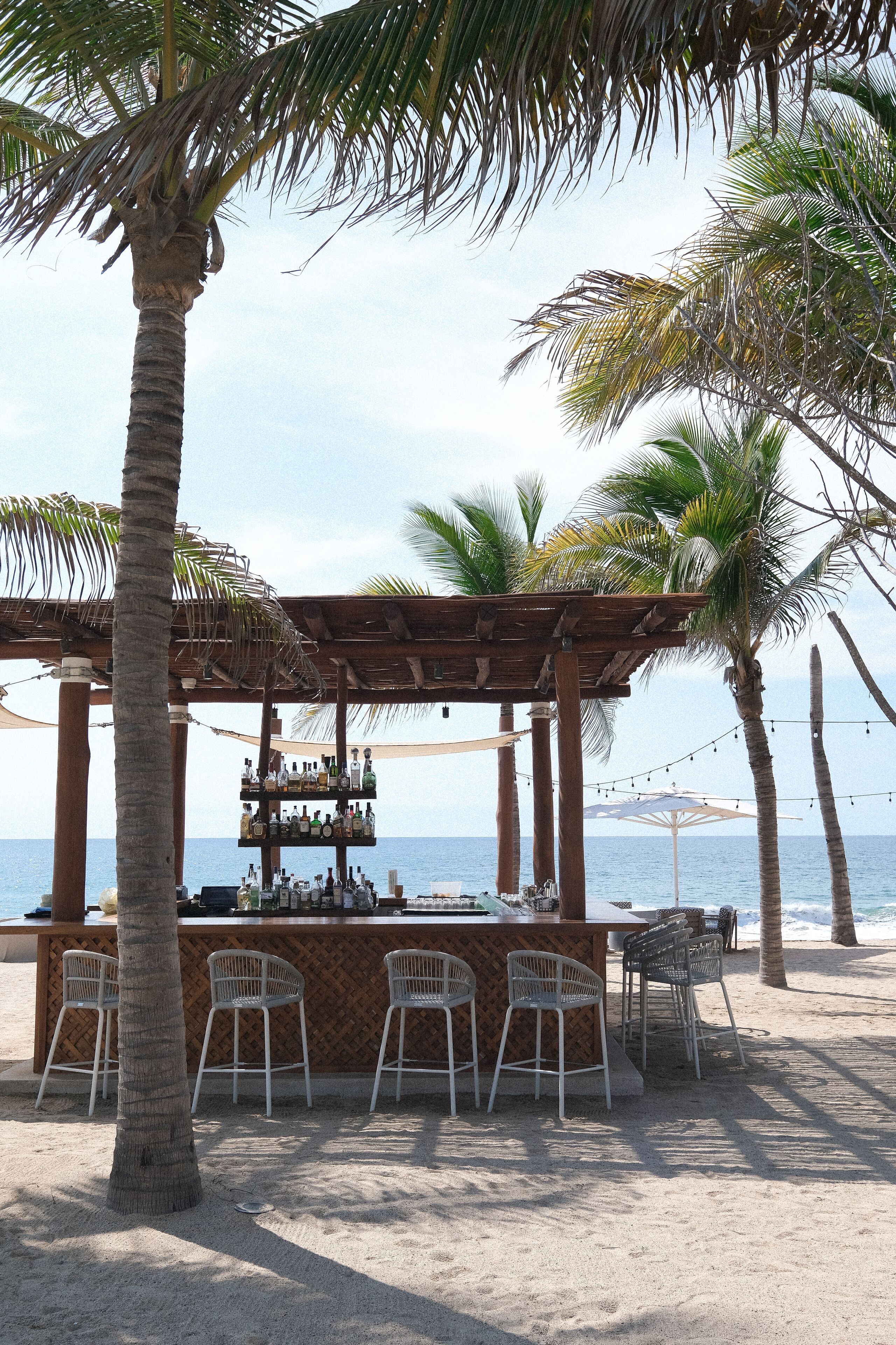 A beach bar with a wooden pergola, white bar stools, and a view of the ocean.