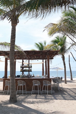 A beach bar with a wooden pergola, white bar stools, and a view of the ocean.