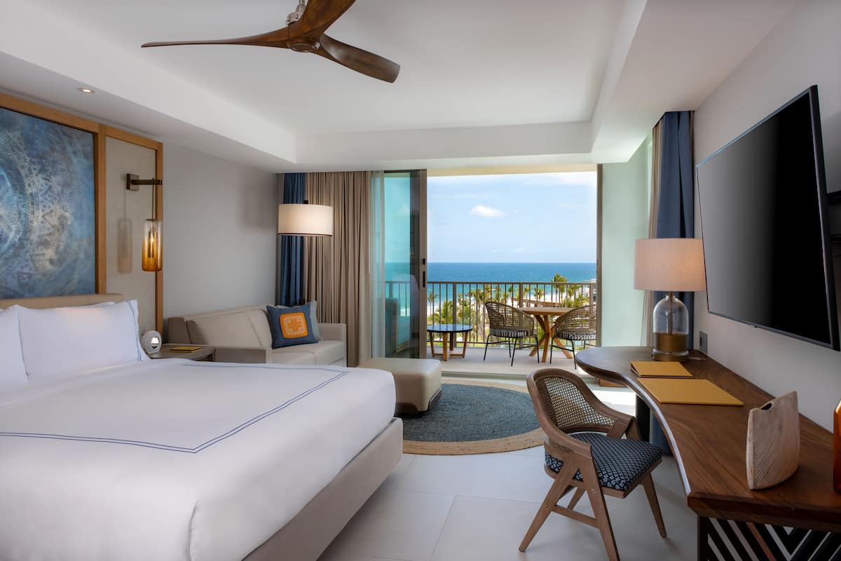 A hotel room with a large bed, a desk with a flat-screen TV, a balcony with a view of the ocean, and a ceiling fan.
