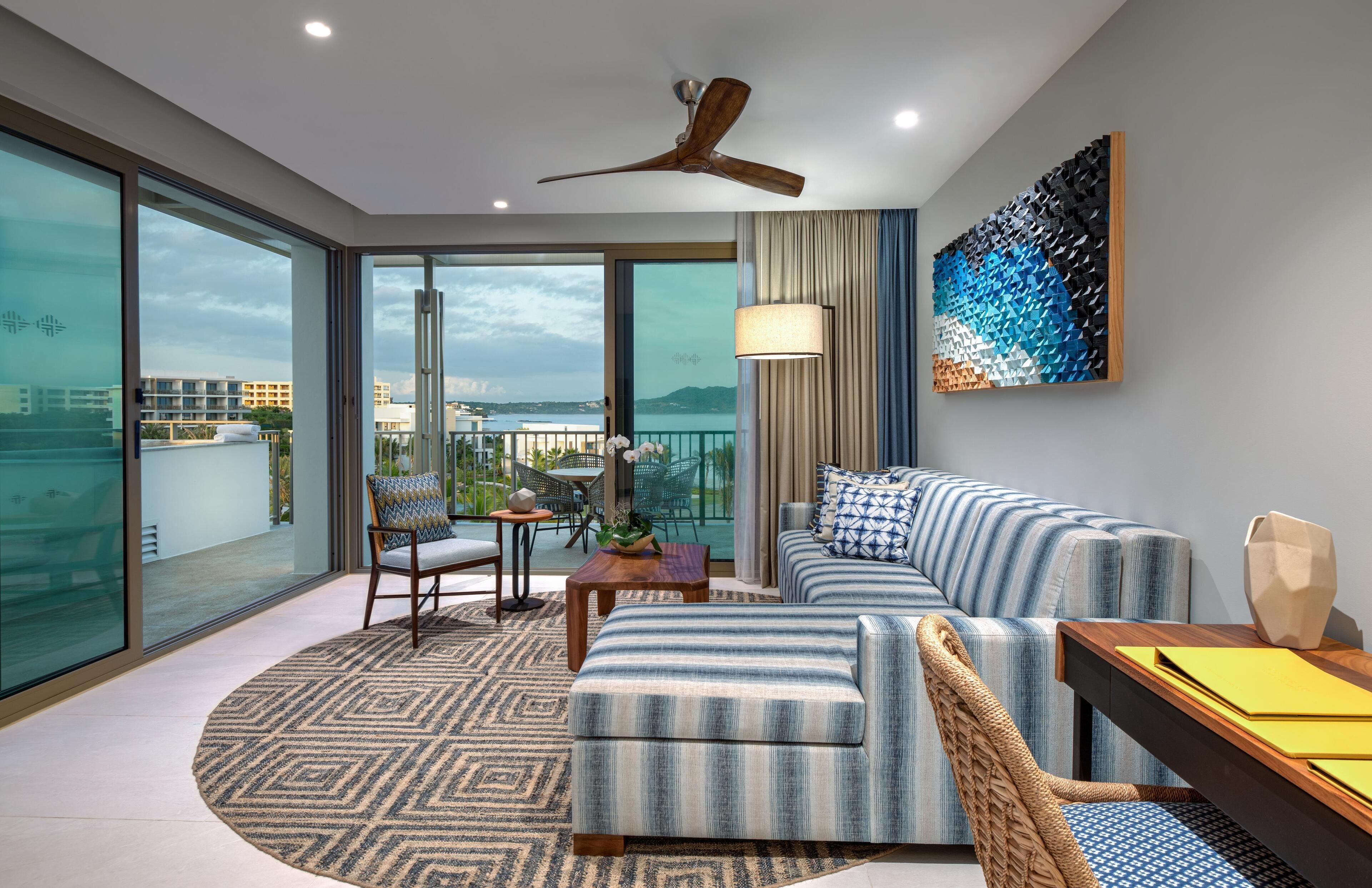 Una sala moderna con sofá, sillón y mesa de centro, un ventilador de techo y vista al mar a través de amplios ventanales.