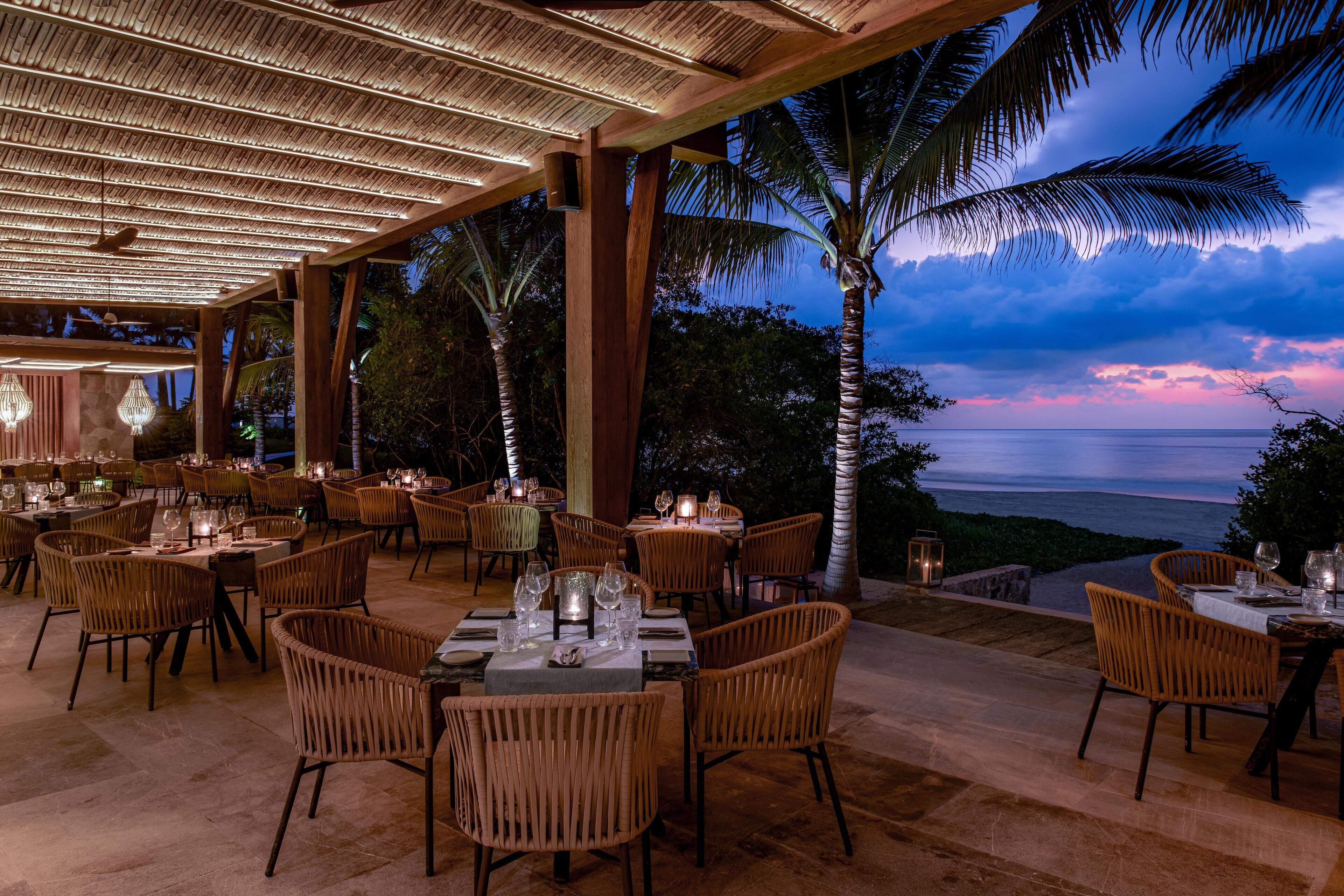 An outdoor dining area with tables and chairs, a thatched roof, and a view of the ocean at dusk.