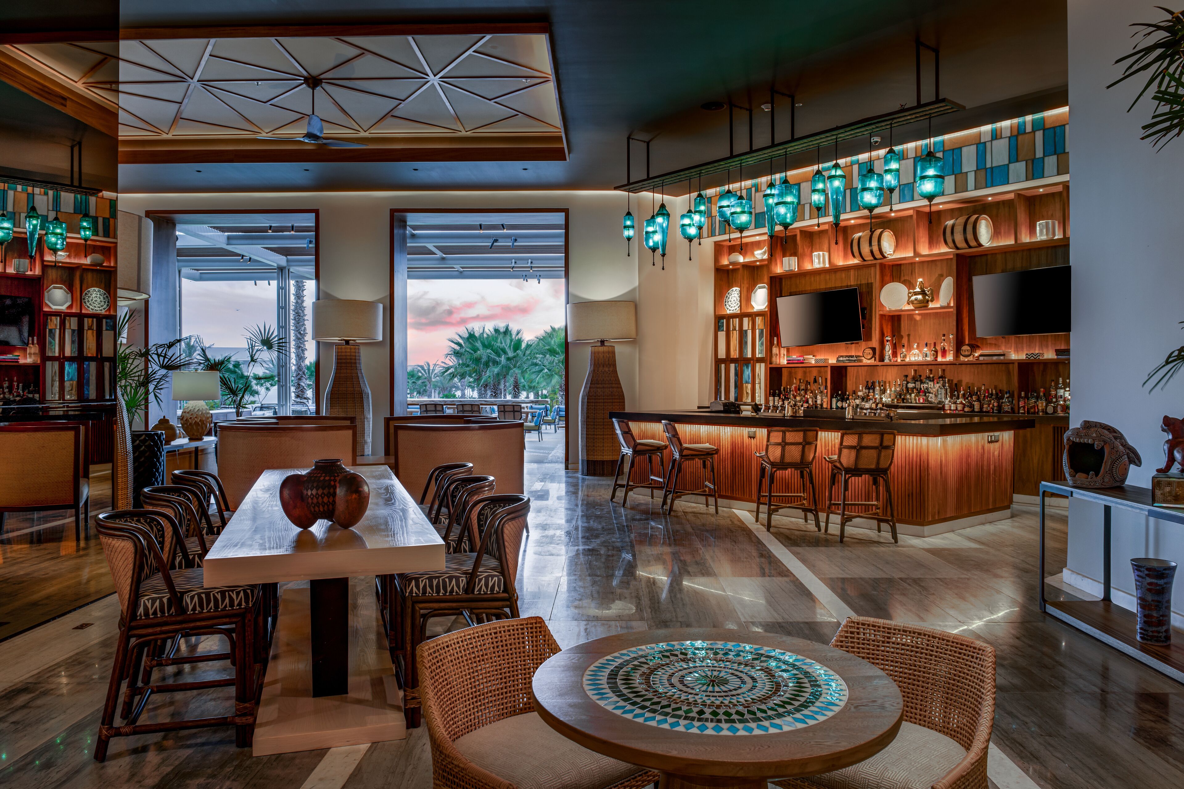 A spacious dining area with a long table and multiple chairs, a bar with stools, and a view of palm trees through large windows.