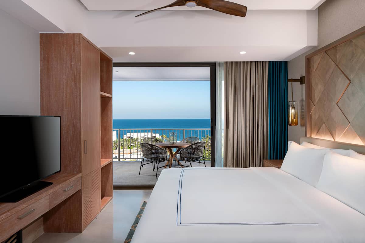 A hotel room with a bed, a TV, a balcony with a view of the sea, and a dining area with chairs.
