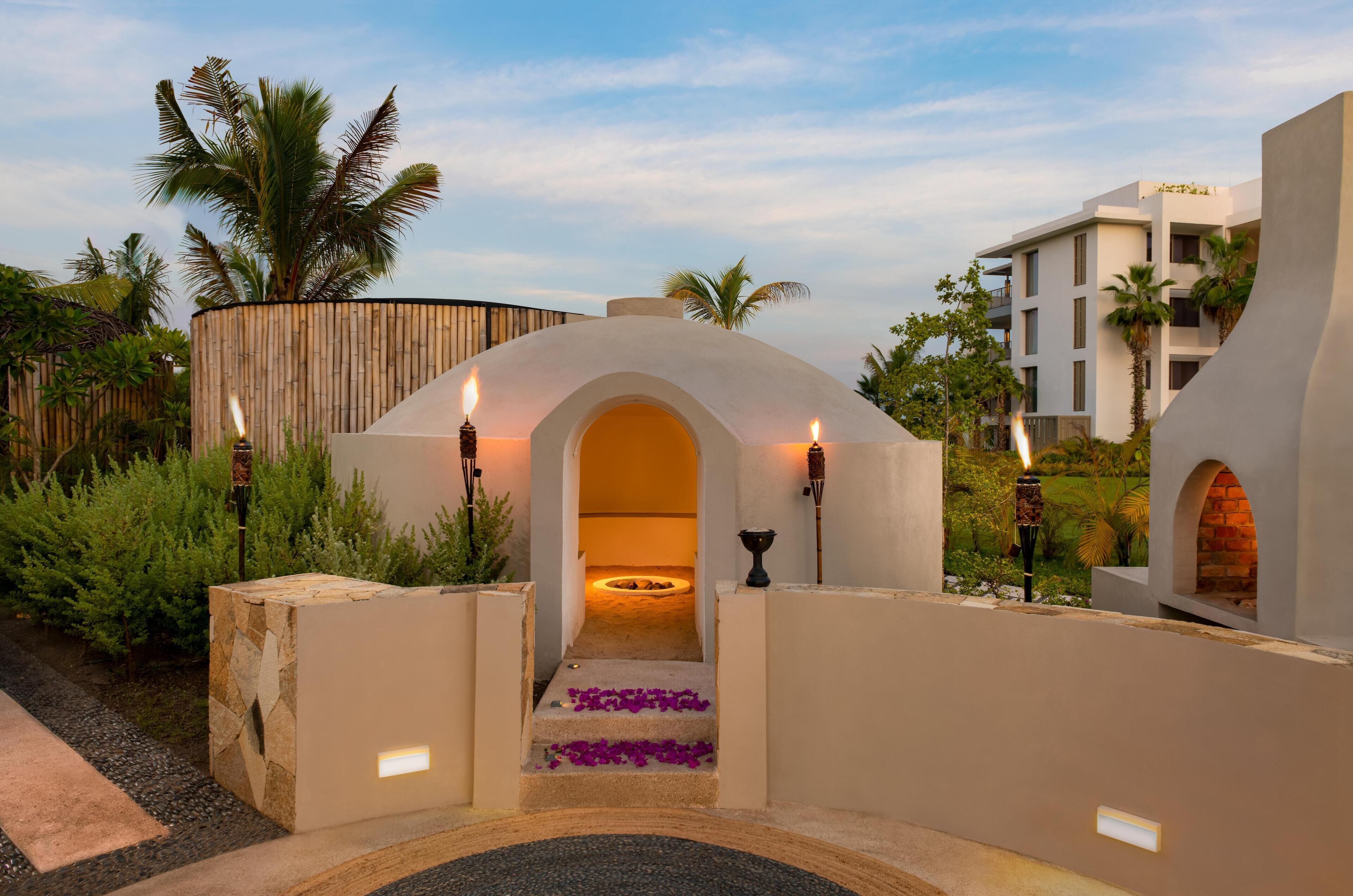 A modern resort entrance with a stone pathway leading to an arched doorway, surrounded by lush greenery and palm trees.