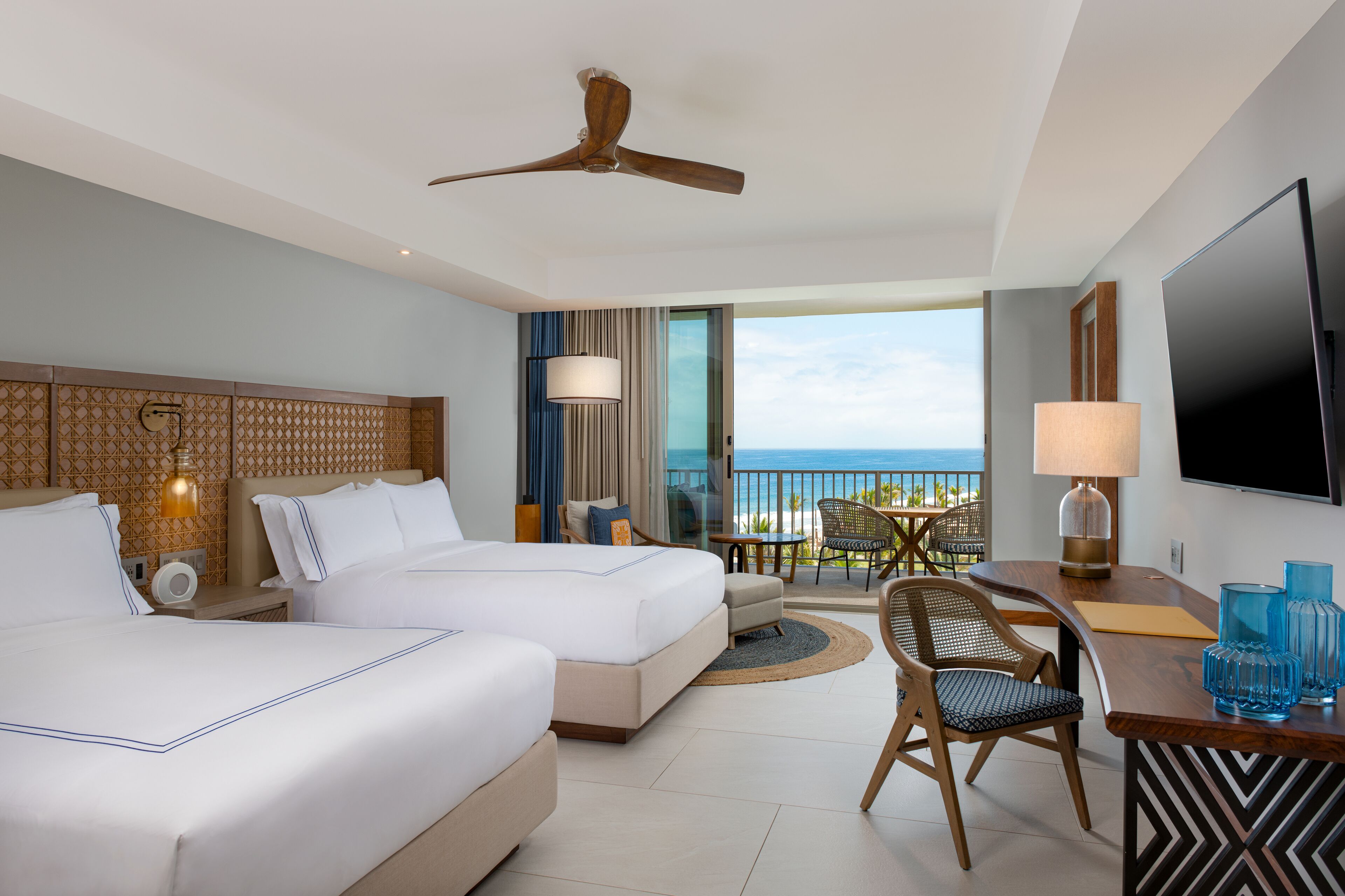 A hotel room with two beds, a balcony with a view of the ocean, a ceiling fan, a TV, and a dining area with a table and chairs.