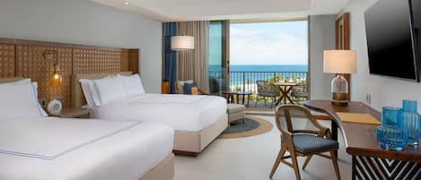 A hotel room with two beds, a balcony with a view of the ocean, a ceiling fan, a TV, and a dining area with a table and chairs.