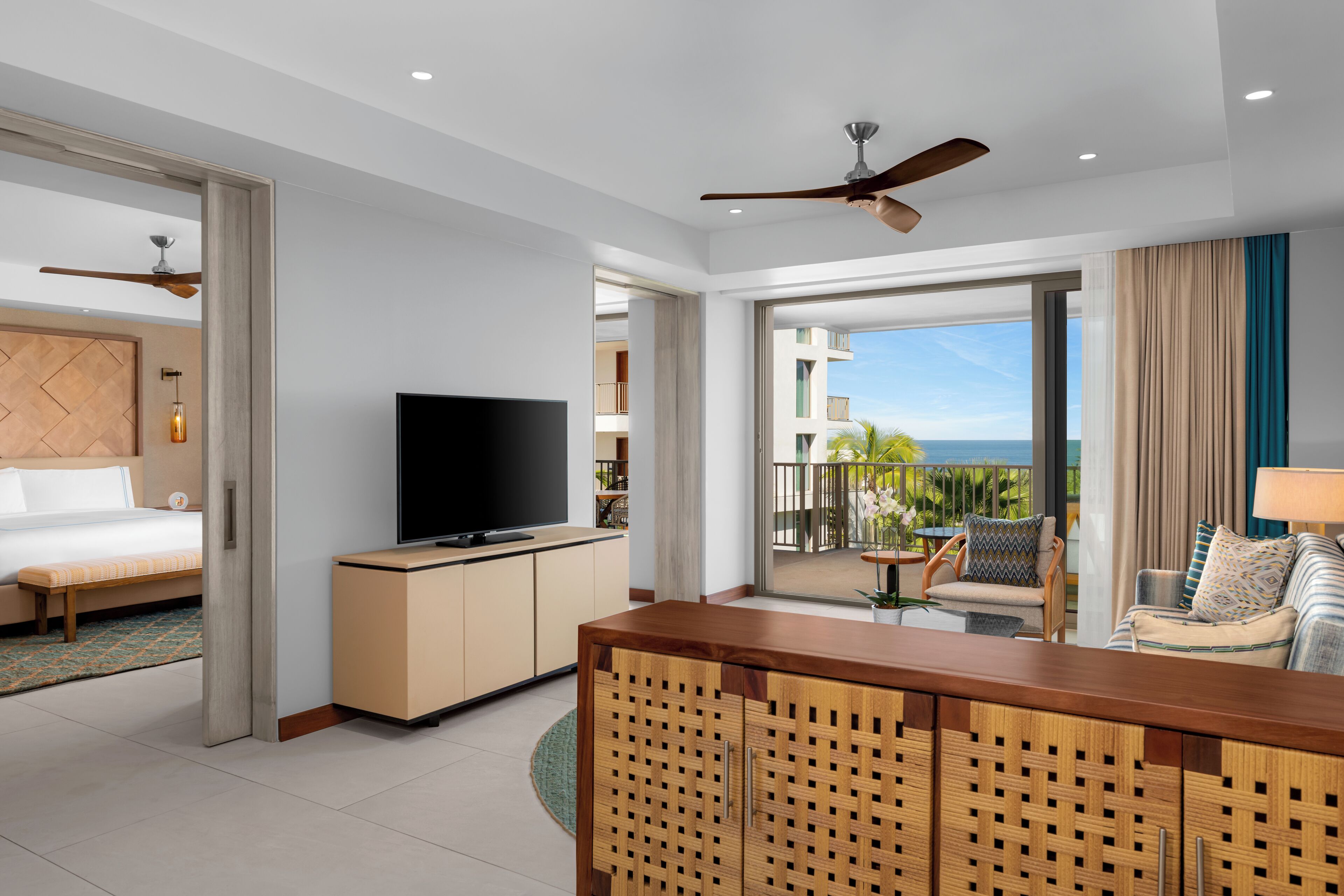A hotel room with a bed, TV, wooden desk, and balcony with a view of the ocean.