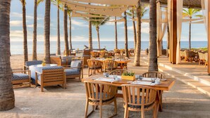 Un restaurante frente al mar con mesas y sillas de madera, luces colgantes y palmeras.