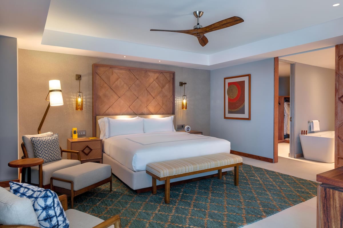 A hotel room with a large bed, a bench, a desk, a chair, a ceiling fan, and a bathroom visible through an open door.