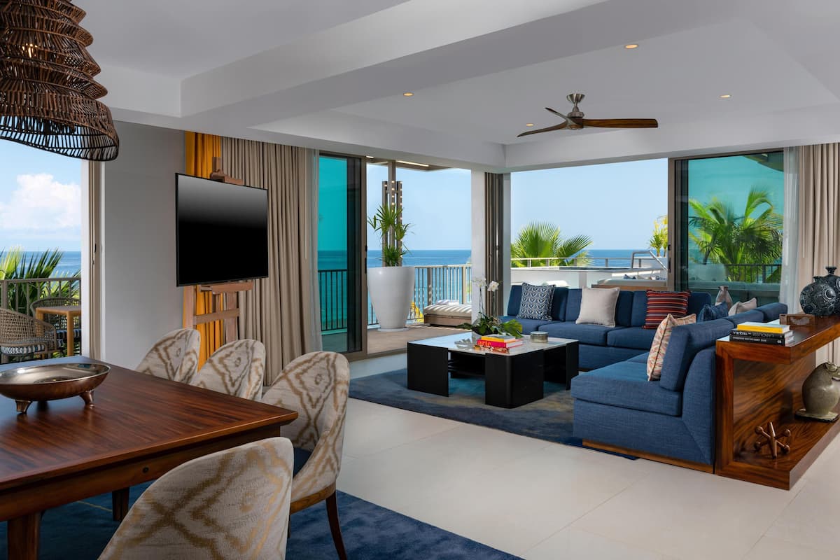 A modern living room with a large TV, comfortable seating, a dining area, and a balcony with a view of the ocean.