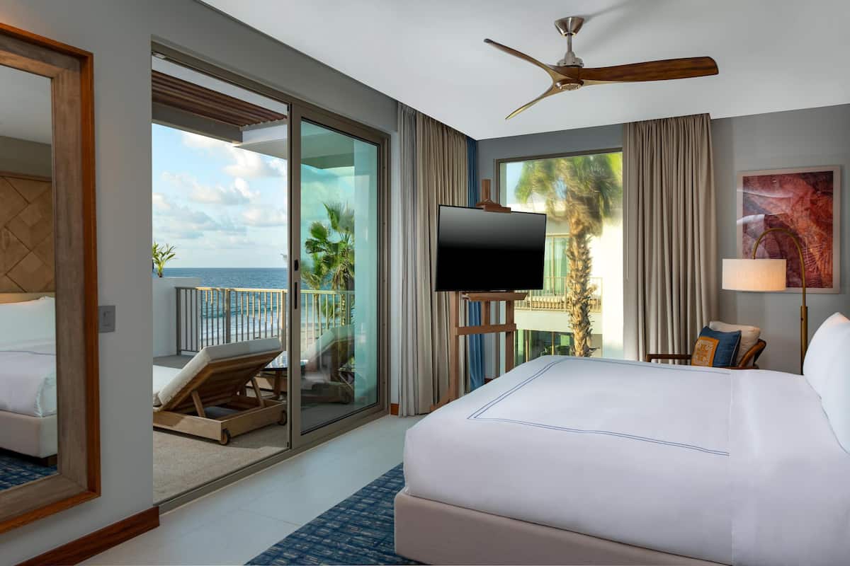 A hotel room with a bed, a TV, a ceiling fan, a balcony with a chair, and a view of the ocean.