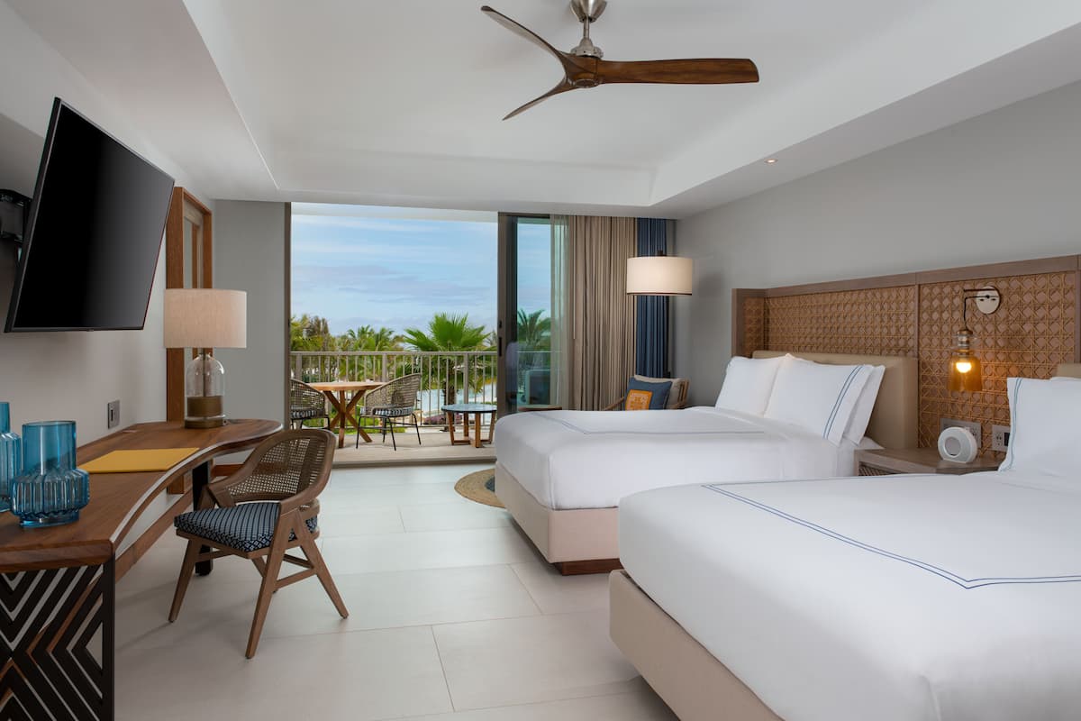 A hotel room with two beds, a desk with a TV, a chair, a balcony with a view, and a ceiling fan.