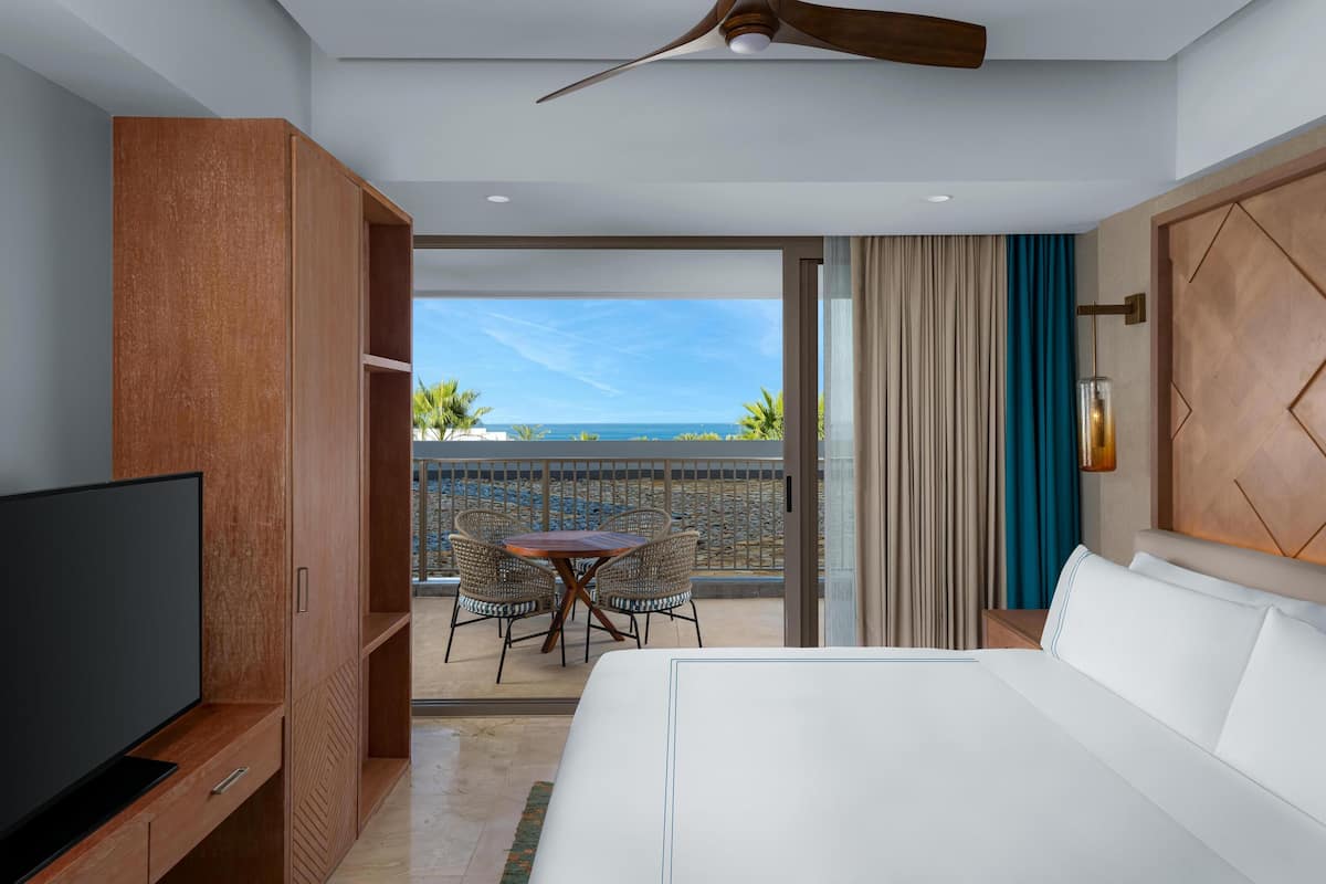 A hotel room with a large bed, a TV, a wooden wardrobe, a balcony with a table and chairs, and a view of the ocean.