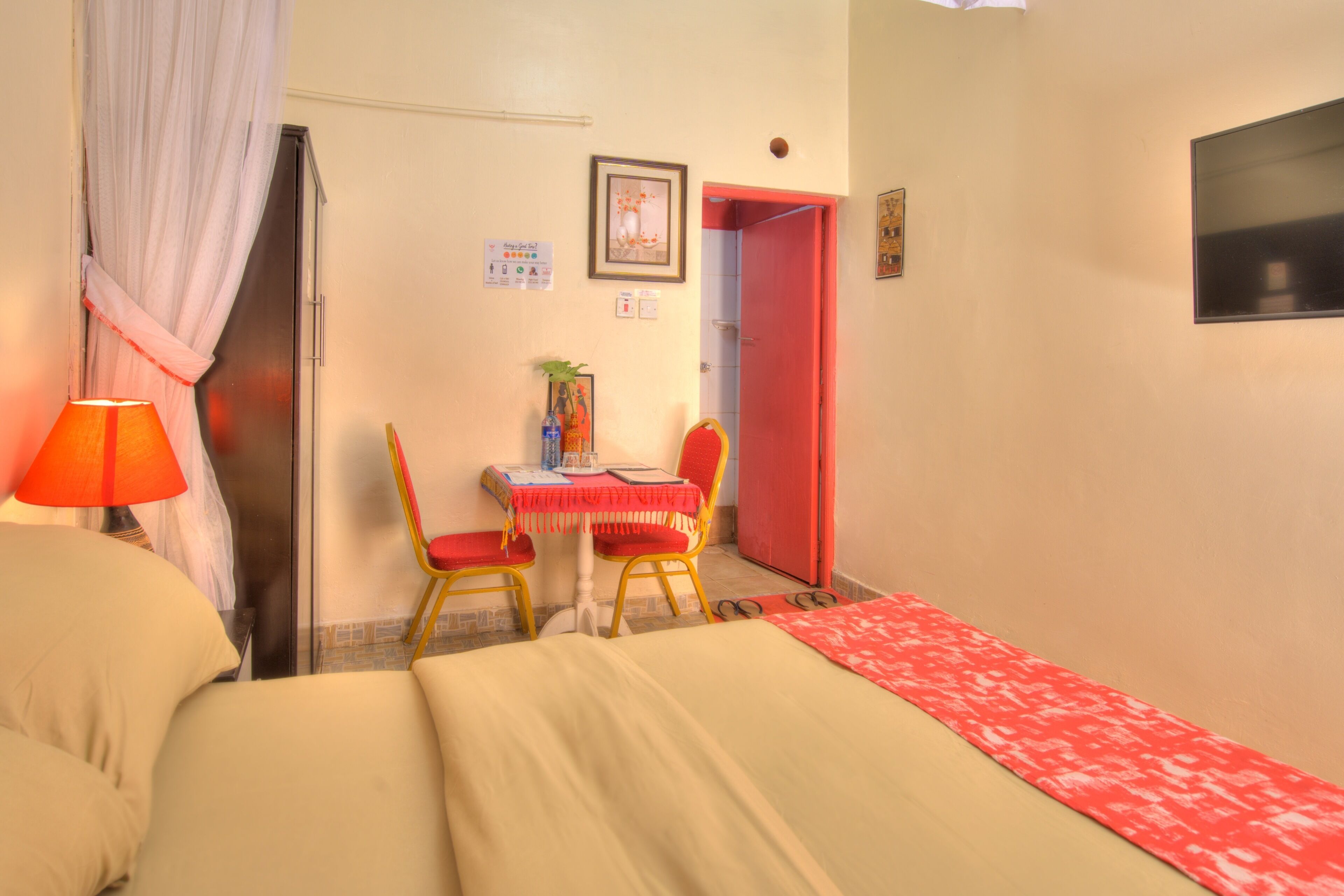 A hotel room with a bed, a desk with a chair, a red door, and a window.