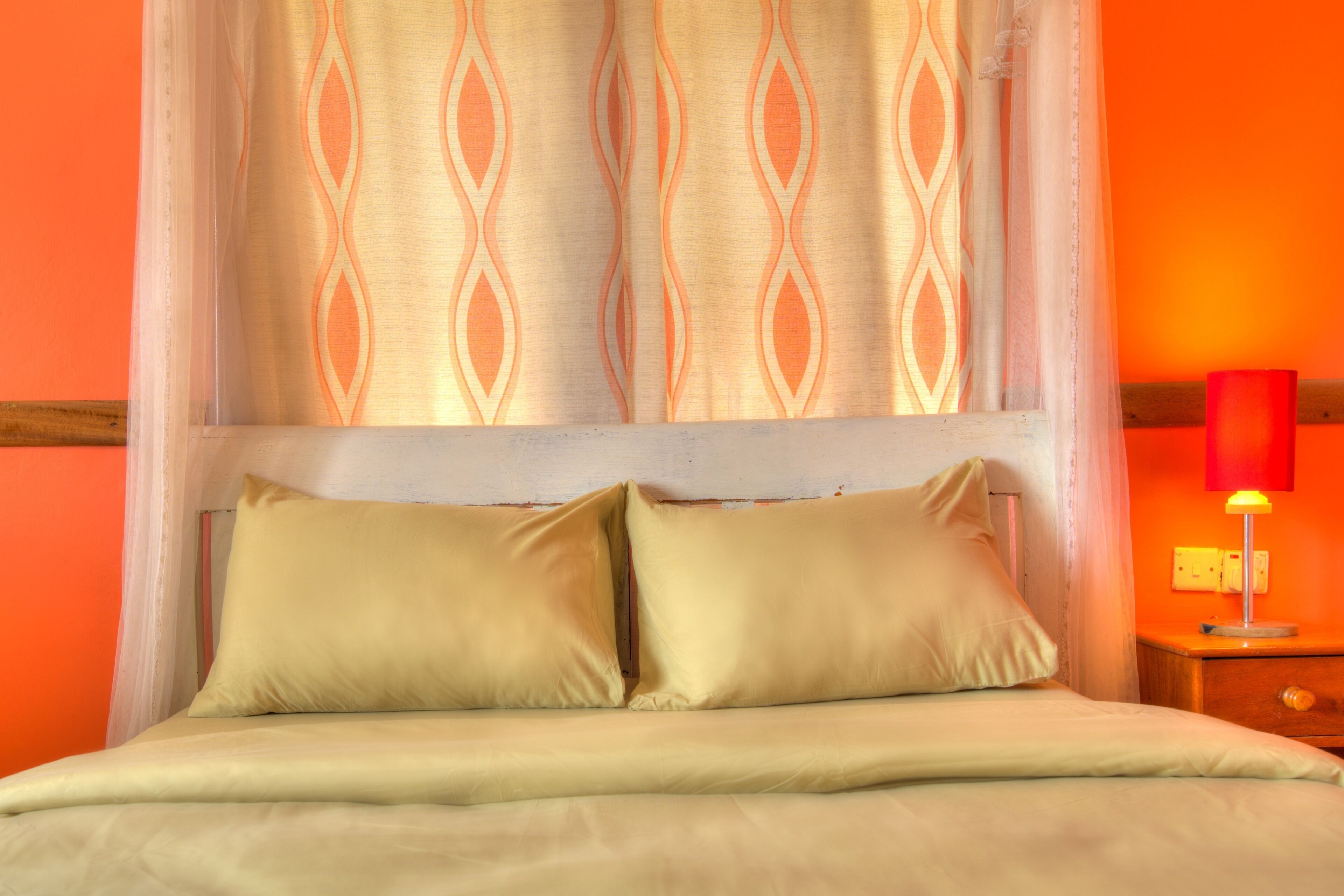 A bed with two pillows, a bedside table with a lamp, and a window with a patterned curtain.