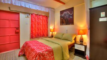A hotel room with a bed, two bedside lamps, a red door, a red curtain, a painting of a buffalo, and a refrigerator.