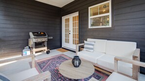 A patio area with a white sofa, a round wooden table, a grill, and a rug.