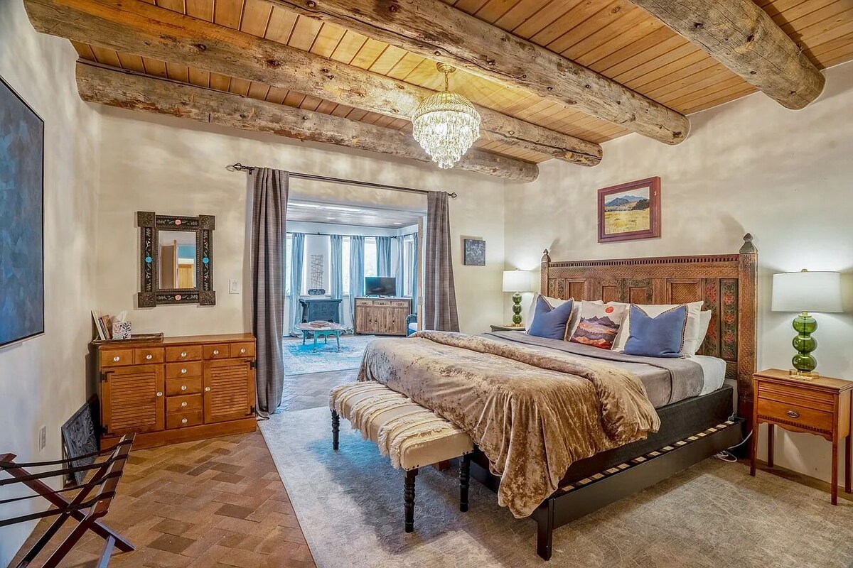 Een ruime slaapkamer met een groot bed, houten balken in het plafond, een kroonluchter en uitzicht op een balkon.