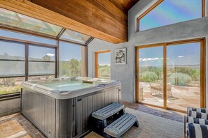 A hot tub in a room with large windows, a wooden ceiling, and a view of a patio area. - Cielo Taos (Arroyo Hondo)