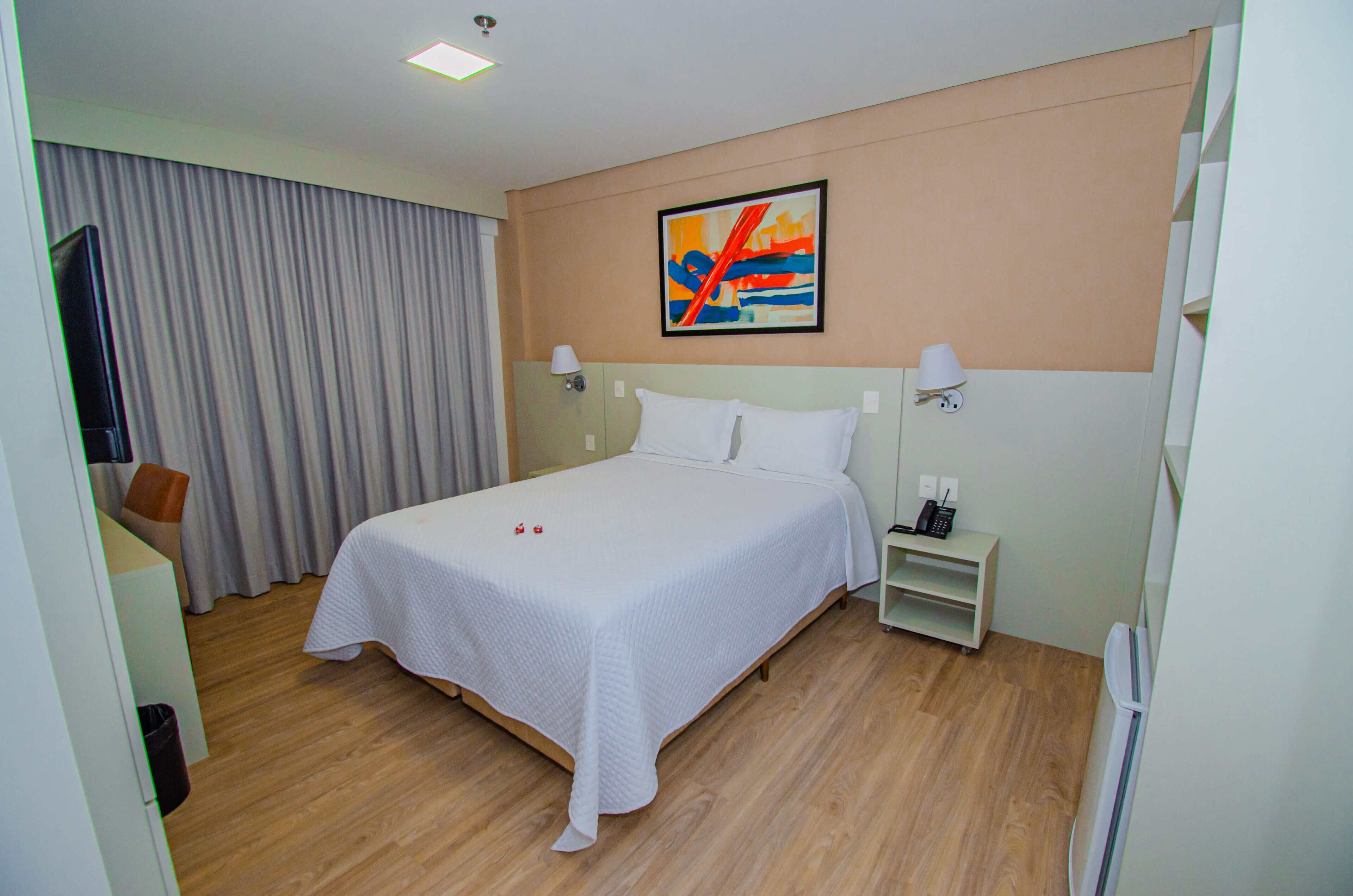 A hotel room with a bed, a desk, a chair, a nightstand, a lamp, a painting, and a window with curtains.