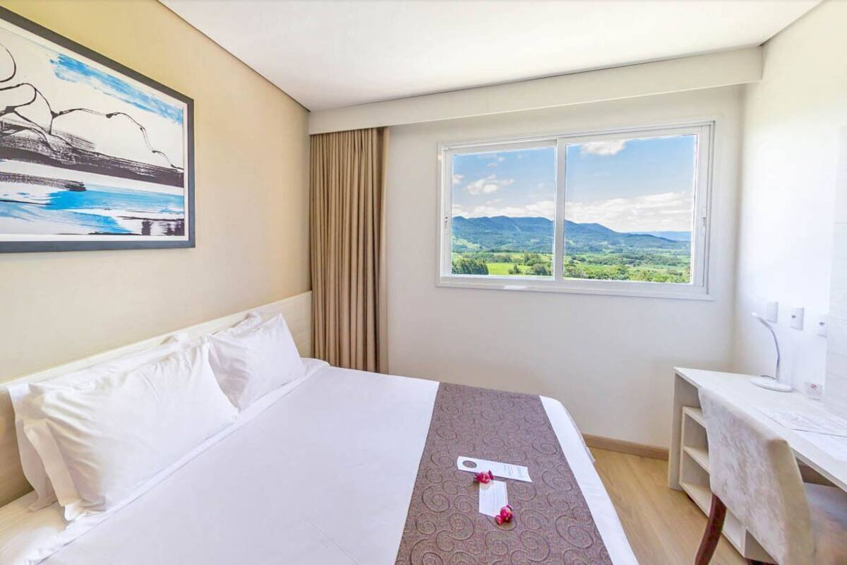 A hotel room with a bed, a desk, a window with a view of mountains, and a painting on the wall.