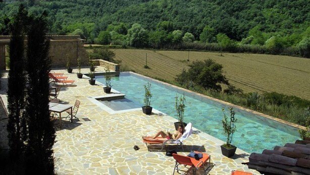 Zona junto a la piscina con sillones de descanso y plantas en macetas, rodeada por un patio de piedra y vistas a una exuberante ladera verde.