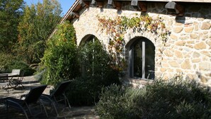 Un edificio de piedra con ventanas arqueadas y techo de tejas, rodeado de exuberante vegetación y mesas al aire libre.