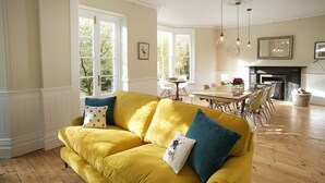 A yellow sofa with blue and white cushions in a well-lit room with wooden floors, large windows, and a dining area with a fireplace. - A substantial six double bedroom property located in the heart of St Agnes. (St Agnes)