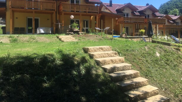 A multi-story building with a red-tiled roof, wooden balconies, and a grassy courtyard with stone steps leading to the entrance.