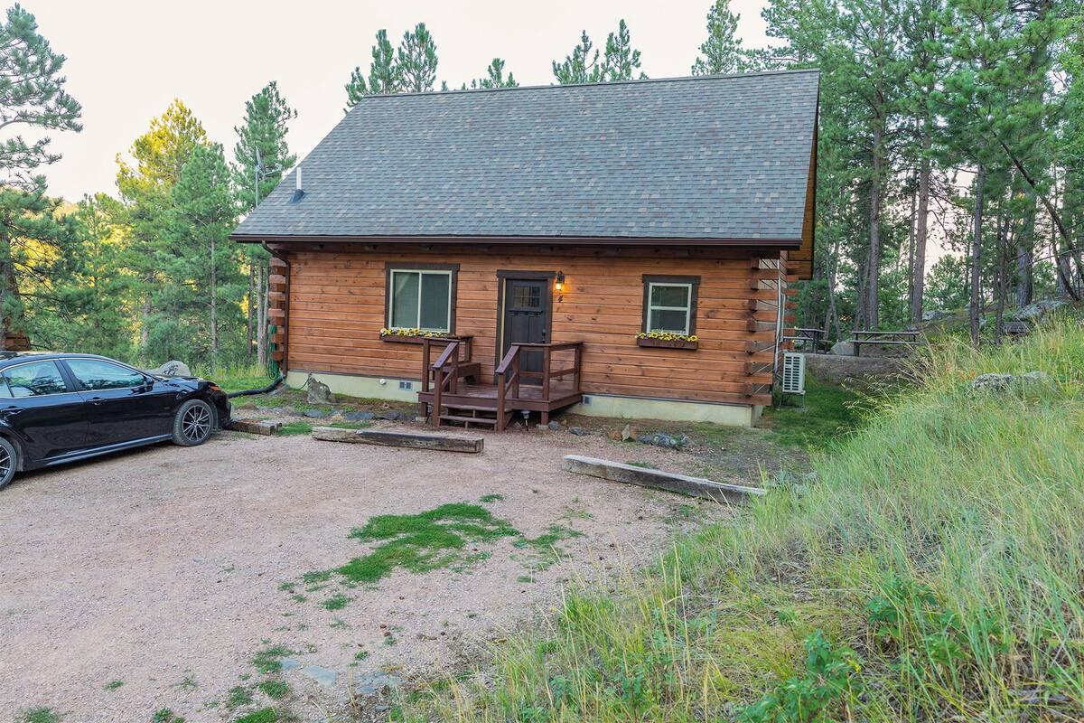 A log cabin with a gravel driveway, a car parked in front, and a picnic table nearby.