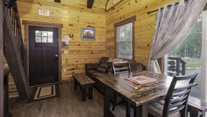 A cabin interior with a wooden interior, a dining table with chairs, a sofa, a window with curtains, and an exit sign. - Freedom Ridge Cabins (Hill City)