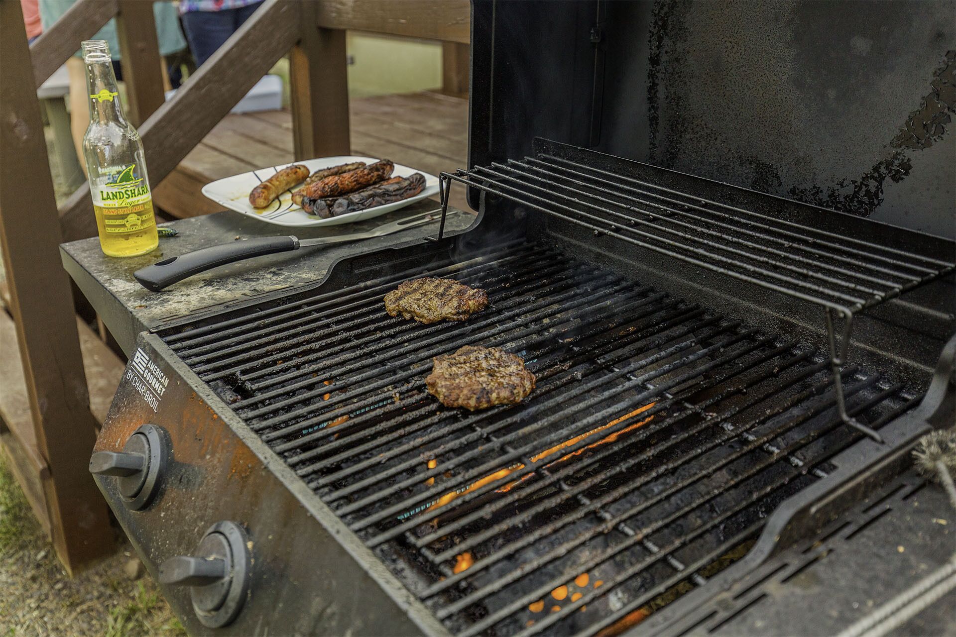 A grill with two burgers cooking, a bottle of beer, and a plate with sausages.