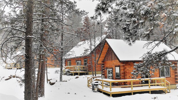A wooden cabin with a covered deck and a snow-covered roof, surrounded by trees. - Freedom Ridge Cabins (Hill City)
