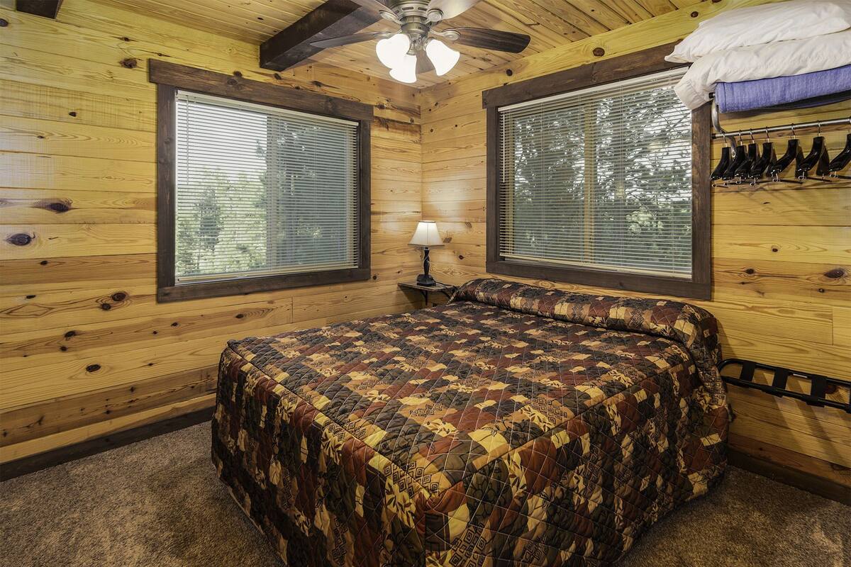 A wooden cabin bedroom with a bed, two windows, a ceiling fan, and a shoe rack.