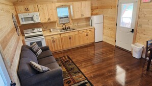 A compact kitchen with wooden cabinets, a microwave, stove, sink, and refrigerator. There's a gray sofa with a patterned rug in the living area. - Freedom Ridge Cabins (Hill City)
