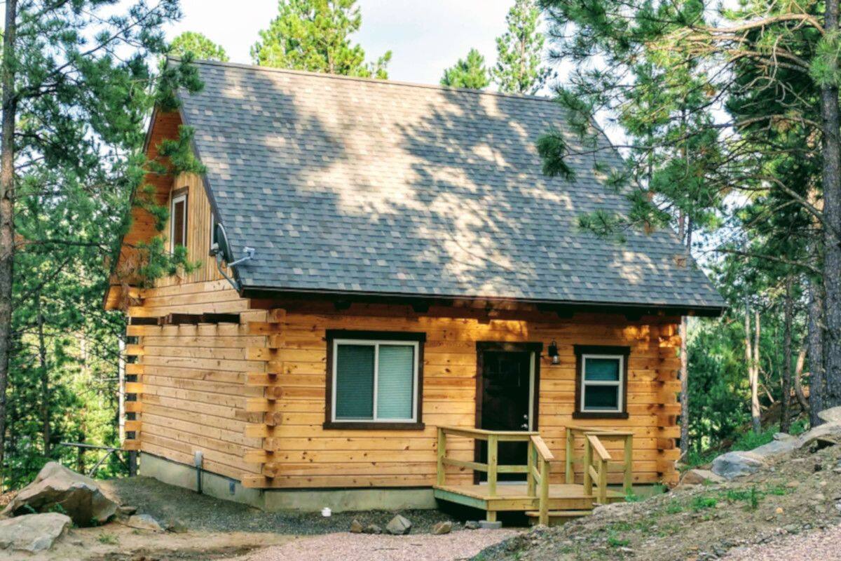A small log cabin with a porch and two windows, surrounded by trees and a gravel path.