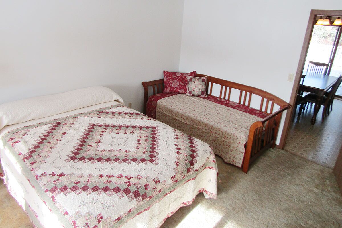 Ein Zimmer mit zwei Betten, eines mit einer Steppdecke und das andere mit einer Blumenbettwäsche. In der Ecke steht ein Holzsessel und ein Tisch.