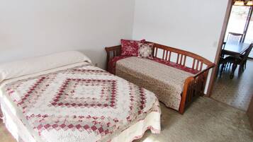 Ein Zimmer mit zwei Betten, eines mit einer Steppdecke und das andere mit einer Blumenbettwäsche. In der Ecke steht ein Holzsessel und ein Tisch.