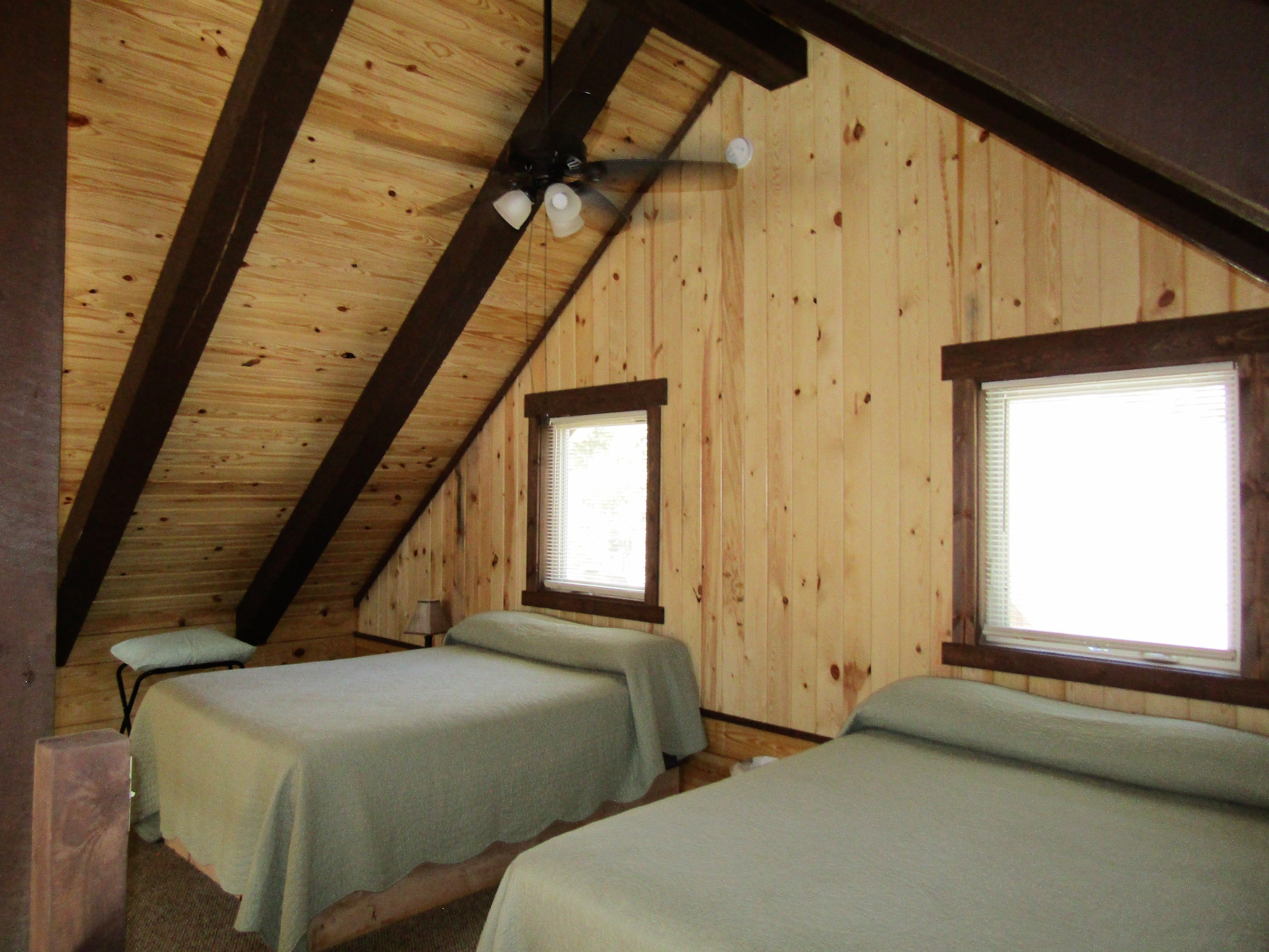 A room with two single beds, wooden walls, a sloped ceiling, and two windows with blinds.
