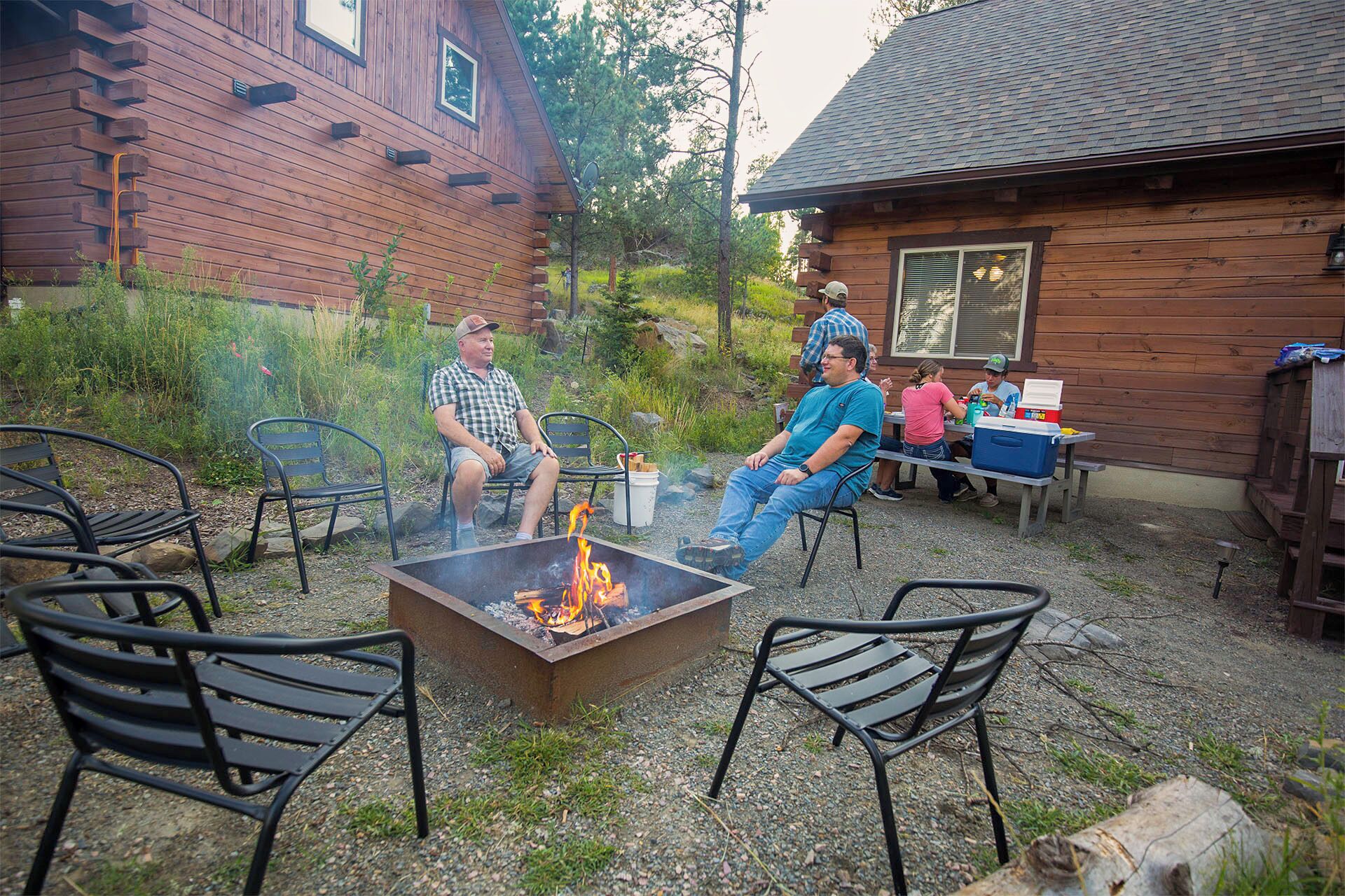 Un espacio al aire libre rústico con sillas de metal y una chimenea, rodeado de cabañas de madera y follaje.
