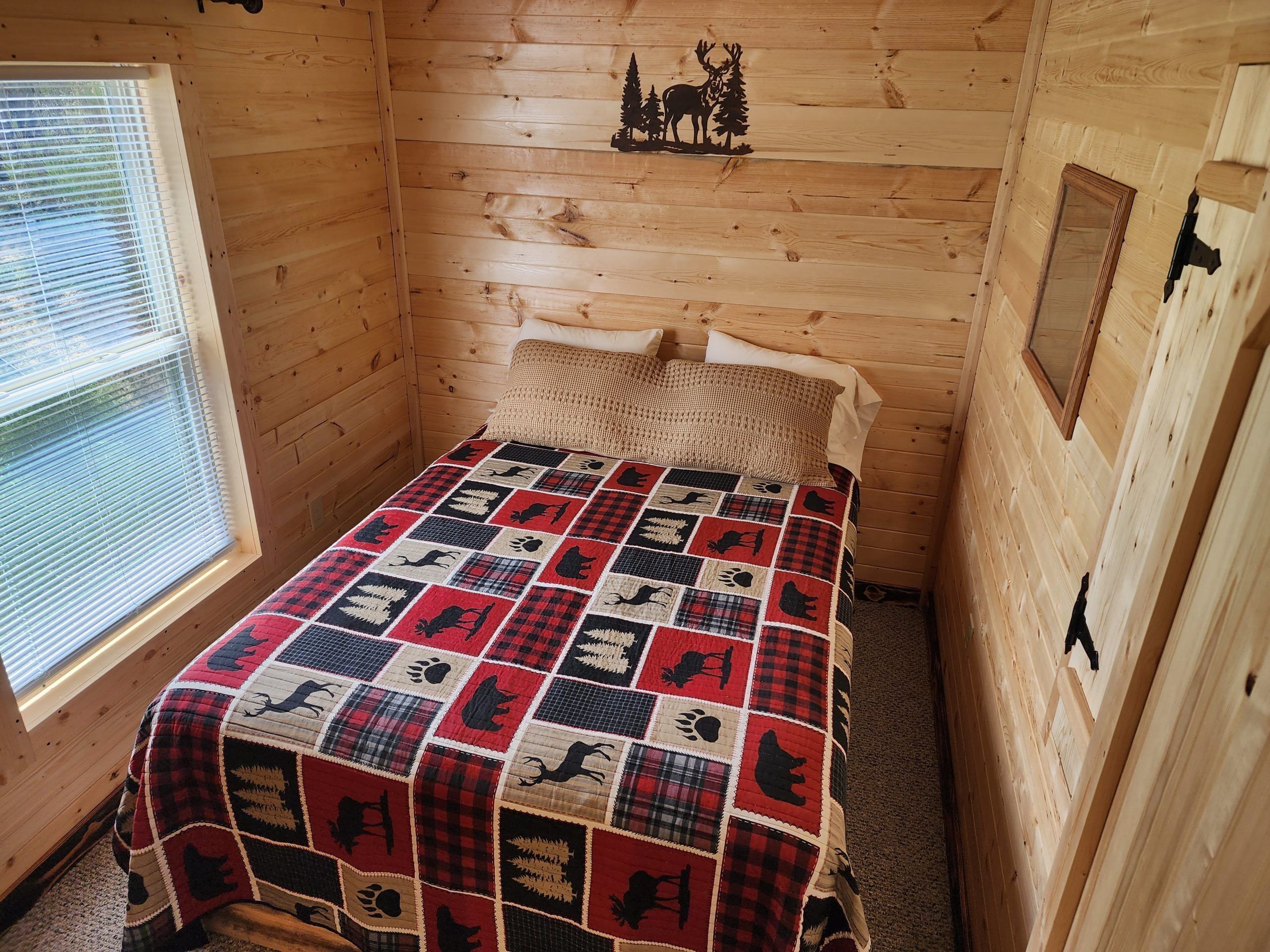 A cozy cabin room with a wooden interior, a quilted bedspread, a window with blinds, and a wall-mounted picture of a deer and trees.