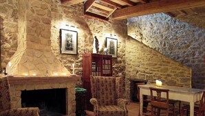 Una habitación con paredes de piedra, una chimenea, una mesa de madera y un sillón con estampado.