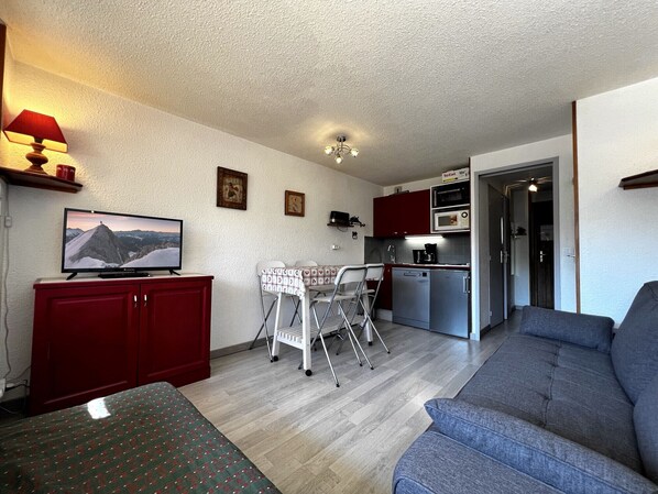 Un espace de vie compact comprenant un canapé, un téléviseur, une table à manger avec des chaises, un coin cuisine et un ventilateur de plafond.