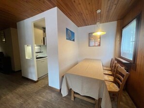A dining area with a table and chairs, a kitchen in the background, and a wooden ceiling.